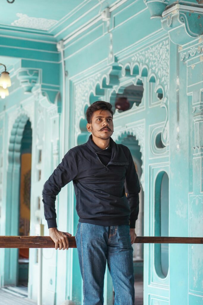 A young man standing indoors against a backdrop of ornate historical architecture with intricate patterns.
